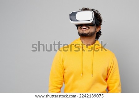 Young gambling fun cool happy smiling Indian man 20s he wearing casual yellow hoody watching in vr headset pc gadget isolated on plain grey color background studio portrait. People lifestyle portrait
