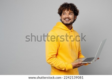 Side profile view young minded smiling happy IT Indian man 20s he wearing casual yellow hoody hold use work on laptop pc computer look aside on area isolated on plain grey background studio portrait Royalty-Free Stock Photo #2242139081