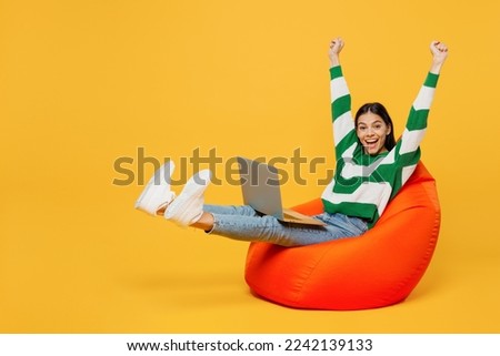 Full body young latin IT woman wears casual cozy green knitted sweater sit in bag chair hold use work on laptop pc computer do winner gesture raise up hands isolated on plain yellow background studio