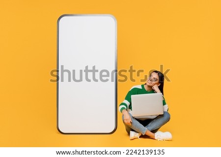 Full body young latin woman wear casual green knitted sweater big huge blank screen mobile cell phone smartphone with area use laptop pc computer isolated on plain yellow background studio portrait