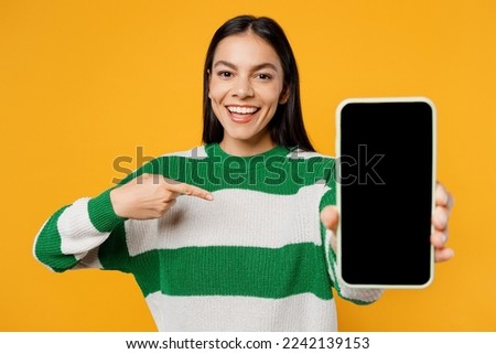 Young brunette latin woman wear casual cozy green knitted sweater hold in hand use point index finger on mobile cell phone with blank screen workspace area isolated on plain yellow background studio