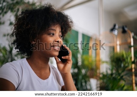 Close-up face of cheerful African-American young woman talking on smartphone, having pleasant phone talk in living room at home with modern green biophilic interior design.
