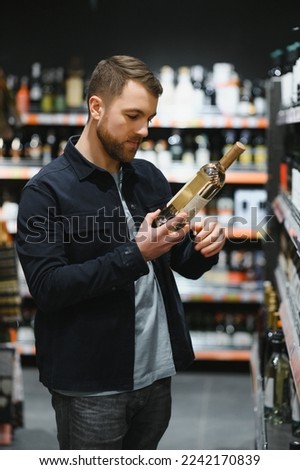 Portrait of young glad positive male customer selecting wine in supermarket. Royalty-Free Stock Photo #2242170839
