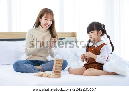 Asian young teenage female mother nanny babysitter in casual sweater and jeans sitting touching little cute preschooler daughter girl head on bed in bedroom while playing brick tower toy game.