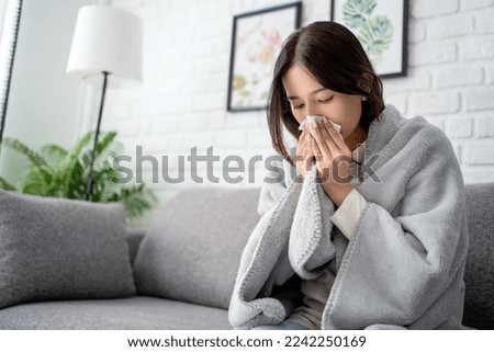 Sick young asian woman sitting under the blanket on sofa and sneeze with tissue paper at home. Royalty-Free Stock Photo #2242250169