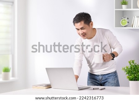 Asian man holding coffee cup and touch laptop on table. Work from home conceptual.