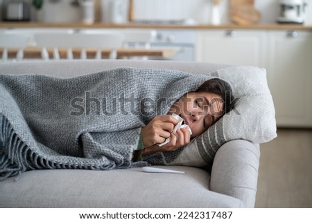 Winter illnesses. Sick female sneezing in tissue while lying under blanket in cold apartment. Young woman suffering from runny or stuffy nose, having cold symptoms. Weakened immunity concept Royalty-Free Stock Photo #2242317487