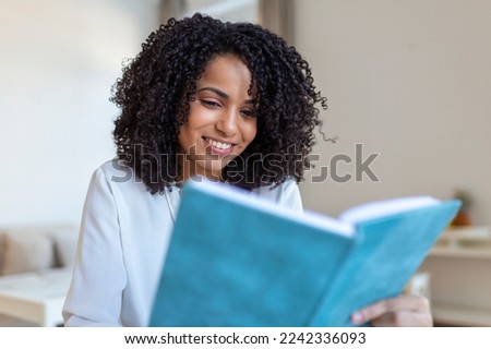 Young beautiful African American woman holding book, University student studying, learning language. leisure, literature and people concept - smiling african american woman reading book at home Royalty-Free Stock Photo #2242336093