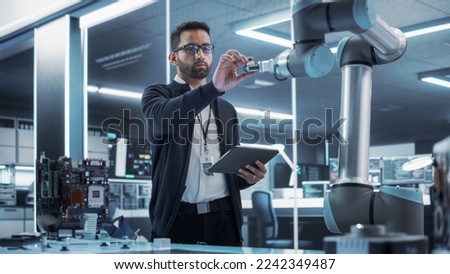 Portrait of a Young Multiethnic Male Engineer Using Tablet Computer. Interacting with a Robotic Arm Machine, Receiving a Microchip from the Hand. High Tech Industrial Laboratory Facility. Royalty-Free Stock Photo #2242349487