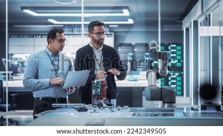 Diverse Colleagues of Industrial Robotics Engineers Experimenting With Robotic Arm. Scientists Use Laptop Computer to Manipulate and Program the Robot to Move and Put Down a Powerful Microchip. Royalty-Free Stock Photo #2242349505