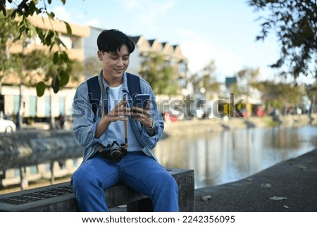 Smiling asian man tourist searching direction on smart phone while sitting on bridge bench near river