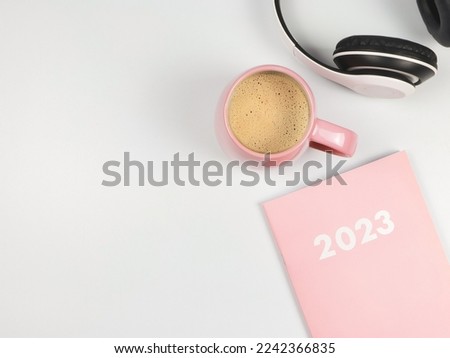 Top view or flat lay of pink diary 2023, headphones and pink cup of coffee on white background.