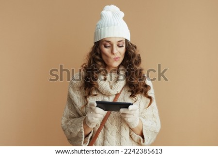Hello winter. pensive trendy woman in beige sweater, mittens and hat on beige background using smartphone applications.