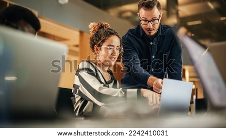 Two Colleagues Working On Laptop in Office. Female Hispanic Help Desk Coordinator Collaborates with White Male Customer Service Agent, They Discuss a Project, Chat, Smile. Teamwork Collaboration Royalty-Free Stock Photo #2242410031