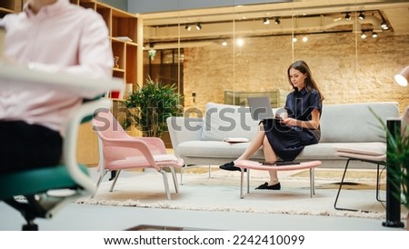 Portrait of White Creative Young Woman Working on a Laptop in Busy Casual Office. Female Team Lead Smiling While Checking her Team Performance Data. Colleagues Walking in Background. Wide Shot Royalty-Free Stock Photo #2242410099
