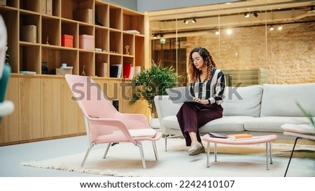 Portrait of Hispanic Creative Young Woman Working on a Laptop in Casual Office. Female Team Lead Smiling While Checking her Team Performance Data. Focused Hard Worker. Wide Shot Royalty-Free Stock Photo #2242410107
