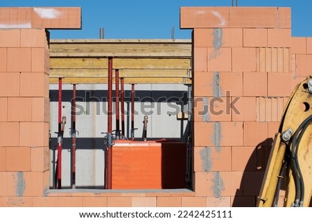 A house is being built on a construction site. The shell has a window opening. Support beams are ready for the ceiling. An excavator stands to the side.