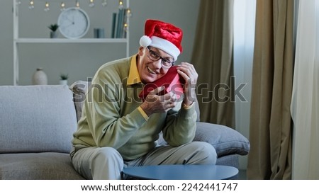 Old Caucasian senior mature grandpa 60s man grandfather male in Christmas X-mas Santa festive red hat at home holding hugging embrace present parcel gift box for New Year celebration looking at camera