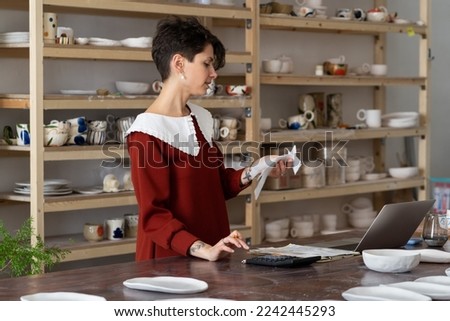 Young busy focused woman entrepreneur doing paperwork, female artisan ceramics shop owner holding receipt using calculator to count expenses, craftswoman running pottery studio, managing finances