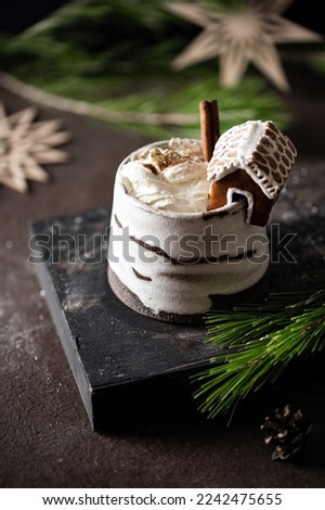 Hot winter drink in a clay mug. Close-up of hot chocolate with mini gingerbread house for single serving. Winter cozy still life with cocoa drink, Christmas decorations, fir tree branch and lights.