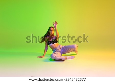 Young sportive girl training, dancing hip-hop over gradient green yellow background in neon light. Youth culture, style and fashion. Concept of dance, youth, hobby, dynamics, movement, action, ad
