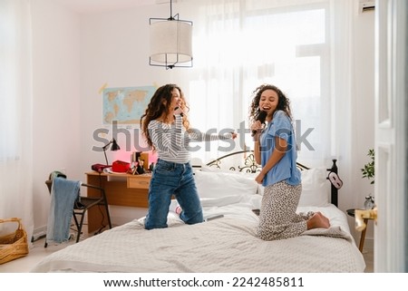 Two cheerful young girls in casual wear holding brushes as microphones and singing songs on bed at home Royalty-Free Stock Photo #2242485811