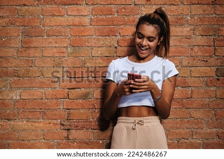 Young woman smiling while using a mobile phone standing outdoors against a brick wall.