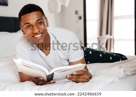 Excited young african man reading book while lying in bed at home Royalty-Free Stock Photo #2242486659