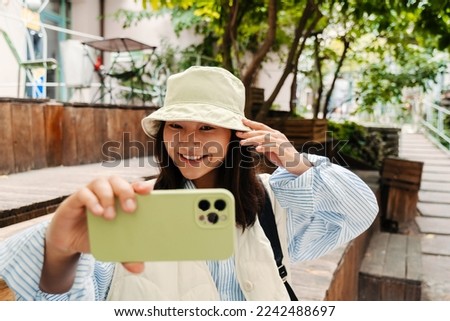 Smiling cute asian girl wearing panama taking selfie with mobile phone while sitting outdoors
