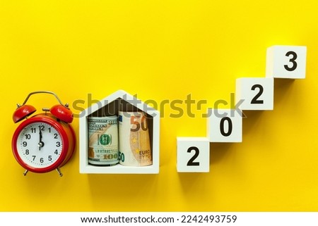Wooden white cubes with numbers 2023 with a house with money and a red alarm clock on a yellow background.Merry Christmas and Happy New Year concept.