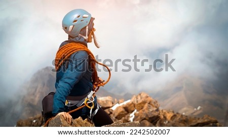 A young cheerful girl in a turquoise helmet, windbreaker and with a red climbing rope sits on the top of the mountain higher than clouds, the traveler successfully climbed the mountains. Royalty-Free Stock Photo #2242502007