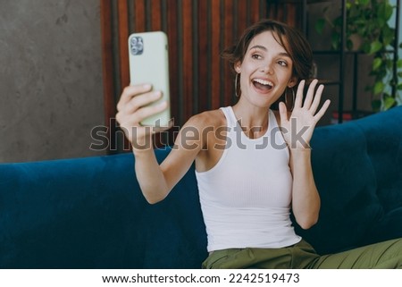 Young smiling happy woman wears white tank shirt do selfie shot on mobile cell phone waving hand sit on blue sofa couch stay at home hotel flat rest relax spend free spare time in living room indoors