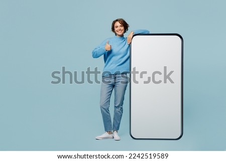 Full body fun young woman wear knitted sweater near big huge blank screen mobile cell phone smartphone with workspace mockup area show thumb up like isolated on plain pastel light blue cyan background