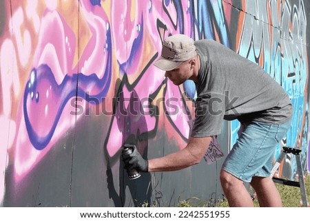 Young caucasian male graffiti artist drawing big street art painting in blue and pink tones. Stylish man in denim shorts and grey t shirt made new graffitti artwork outdoors in bright sunny day