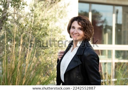 Portrait of positive woman 45+ years old. Outside, building, trees in background. Business woman in black leather jacket, white blouse. Career, business, woman, health care concept. Lifestyle. Royalty-Free Stock Photo #2242552887