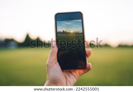Faceless person taking photo of green grassy field under picturesque sky in sunset in summer on smartphone in selective focus