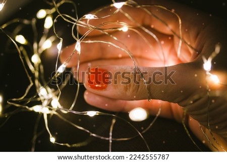 Female hand holding a tangled led garland, glowing lights. Woman preparing to decorate a Christmas tree for New Year's Eve 2023. Thin fingers with orange-red manicure holding a wire on dark background