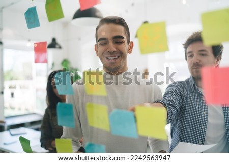 View through glass of male employee showing information on sticky note pointing with marker to coworkers while gathering in modern office creating new strategy