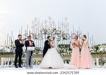 Front view of wedding ceremony outdoors, man in suits kissing woman in white puffy dress, while standing with bridesmaids and groomsmen near floral arch. Royalty-Free Stock Photo #2242575789