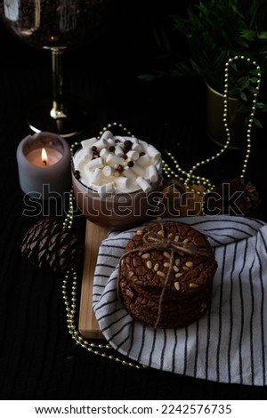 Chocolate cookies with pine nuts and a cup of hot chocolate with marshmallows on white towel and wooden desk. black background. Dark and moody stock photography.