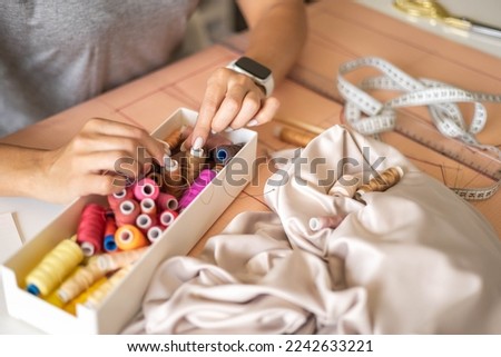 Woman dressmaker hands choosing thread color for sewing beige fabric clothes at workshop studio closeup. Female tailor fashion designer art work modist dressmaking professional occupation top view Royalty-Free Stock Photo #2242633221