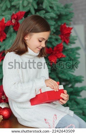 Happy interested pretty little girl opening Christmas gift. Child sitting on a New Year decorated kitchen and holding Xmas present