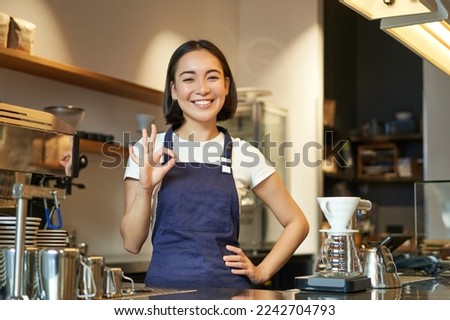 Portrait of smiling korean barista, girl at the counter, wears blue apron, works in coffee shop, shows okay sign. People at work