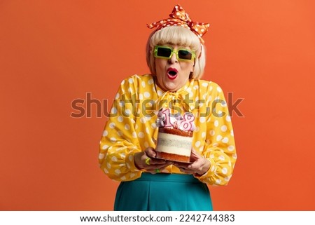 Surprised Grandmother Celebrating Birthday. Amused Fashionable Senior Lady Celebrates Birthday Holds Cake With Burning Candles Isolated Over Orange Background. Positive Granny Celebrates Bday 