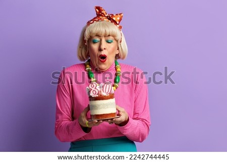Happy Grandmother Blowing Candles. Amused Fashionable Senior Lady Celebrates Birthday Holding Cake With Burning Candles Isolated Over Violet Background. Positive Granny Celebrates Bday 