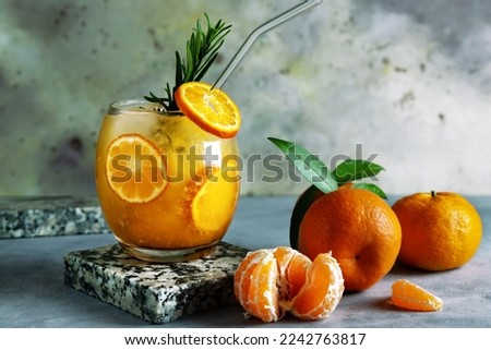 Fresh tasty citrus juice in glass and ripe tangerines on table. Cold summer drink with citrus slices, ice and rosemary branch on grey rustic background. Close up. Selective focus. Blurred background.