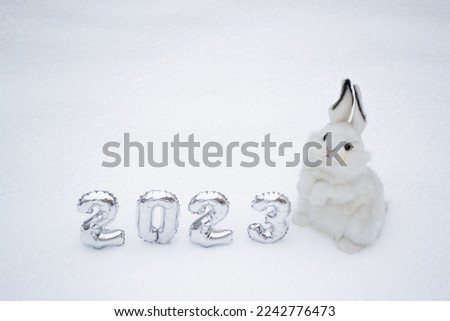 White fluffy bunny toy with silver numbers of the New Year 2023 on the white snow background. Copy space. Symbol of Chinese New Year 2023 