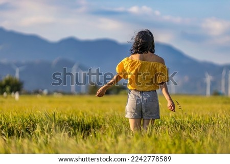 WOMAN RELAXING ON RICE FIELD AND WIND FARM OR WIND PARK BACKGROUND, WITH HIGH WIND TURBINES FOR GENERATION ELECTRICITY WITH COPY SPACE, WIND POWER HAS EQUIPMENT FROM EUROPE. NINH THUAN, VIETNAM Royalty-Free Stock Photo #2242778589