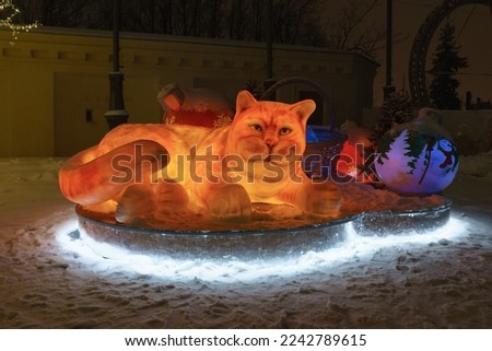 Funny red artificial cat as decoration to the New Year holidays on the city street. Looks at camera. Moscow festive mood. Winter snowy night