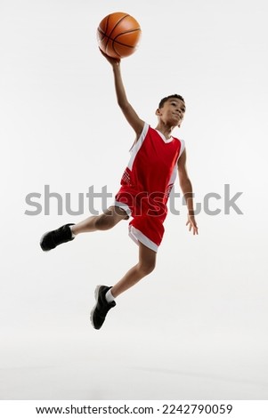 Portrait of boy in red uniform training, playing basketball, throwing ball in a jump on grey studio background. Concept of energy, professional sport, motion, action, hobby, competition, achievement.